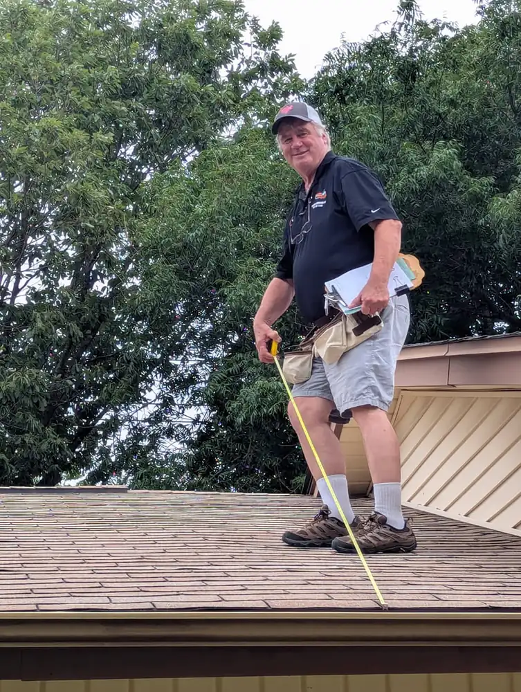 Frank Ruddy standing on a shingle roof with a tape measure and tool belt