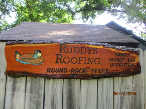 The painted cedar sign mounted under a shingled eave against a board fence