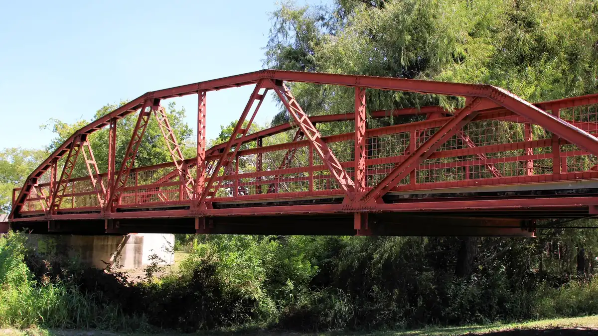 A red steel truss bridge crossing a wooded creek in Fritz Park in Hutto
