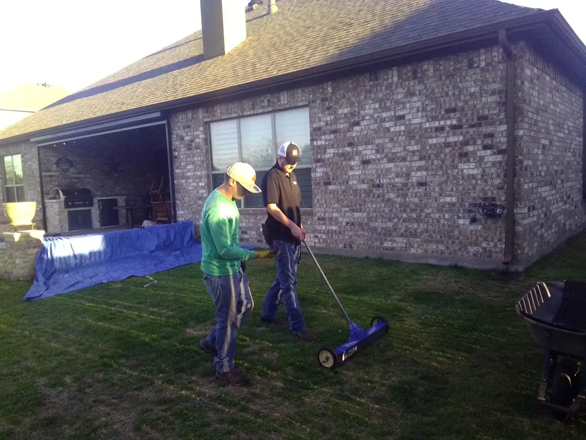 Jake rolling a blue yard magnet across a back lawn after a roof replacement, a blue tarp still along the patio